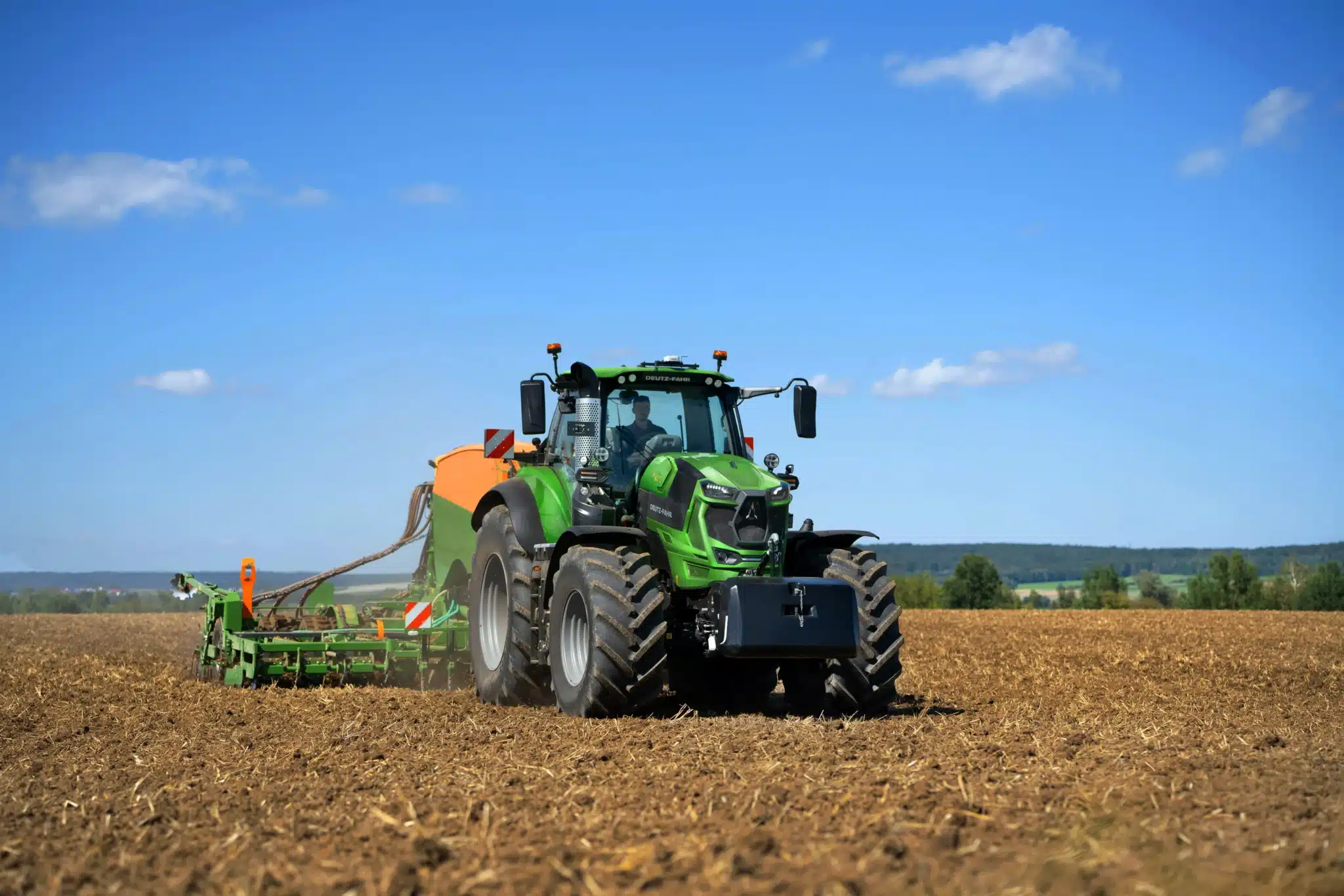 DEUTZ-FAHR 8-Series traktor i grøn farve arbejder på en stubmark med en stor såmaskine eller jordbearbejdningsmaskine i klart solskin.