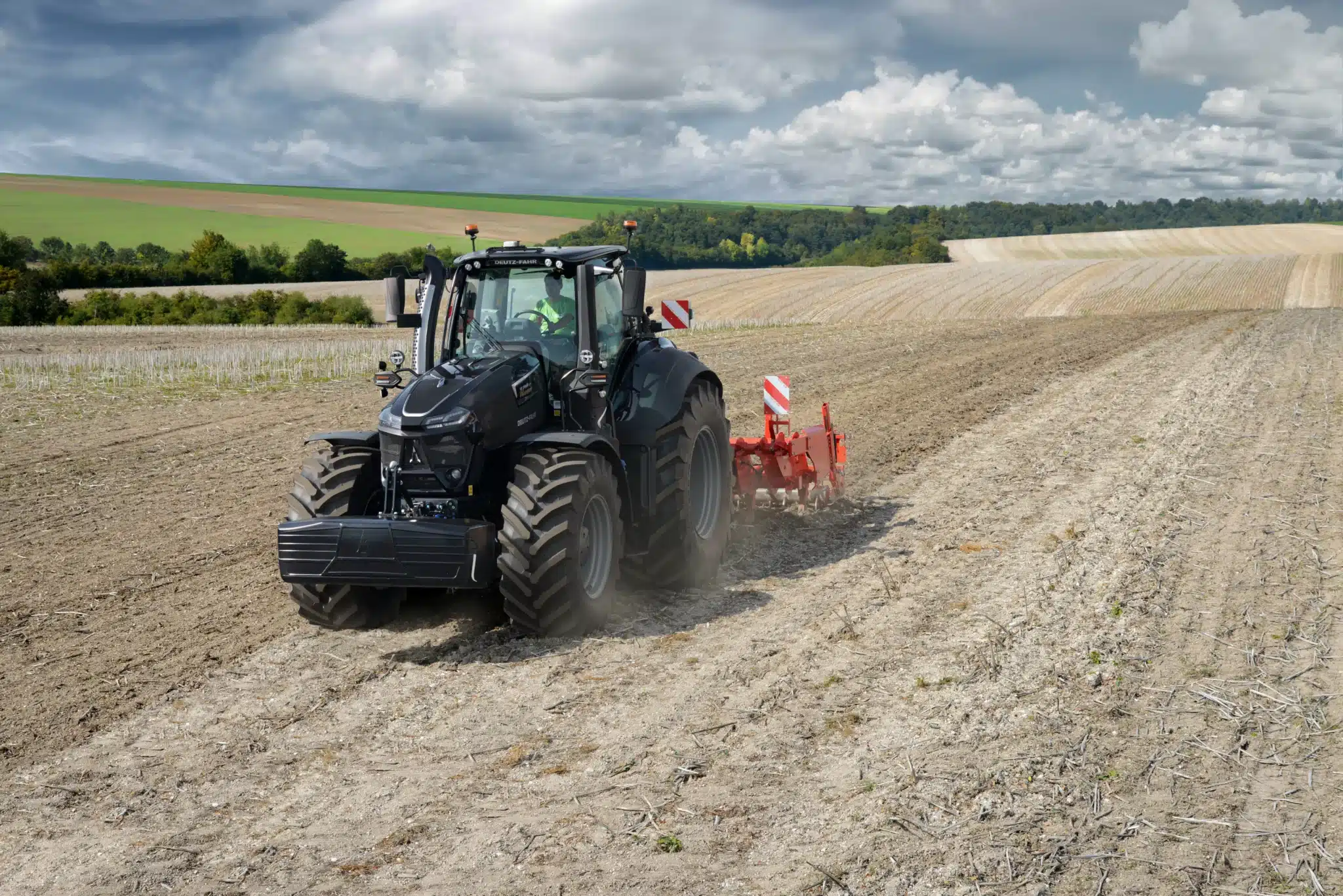 Sort DEUTZ-FAHR Warrior traktor pløjer en stor mark med en rød plov under en dramatisk himmel.